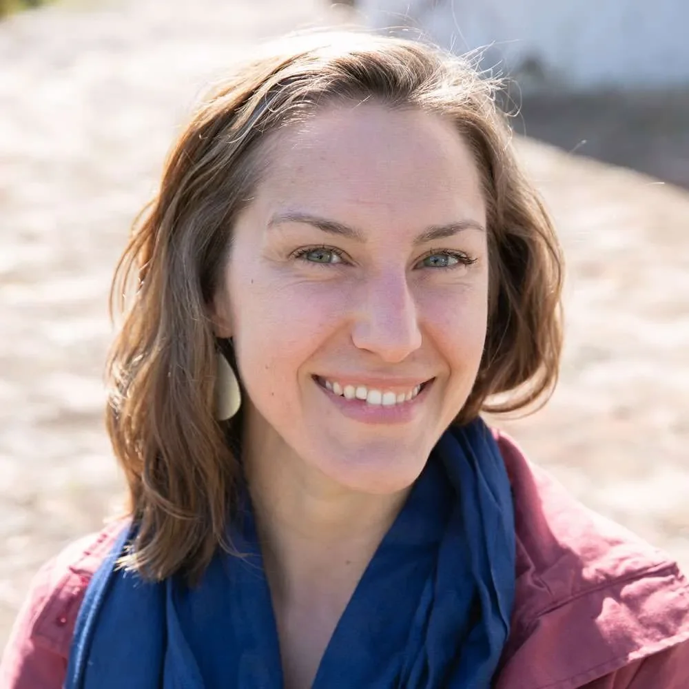 Trisha Mitchner Certified High Performance Coach™ A smiling woman with shoulder-length brown hair wearing a blue scarf and pink jacket outdoors near water.