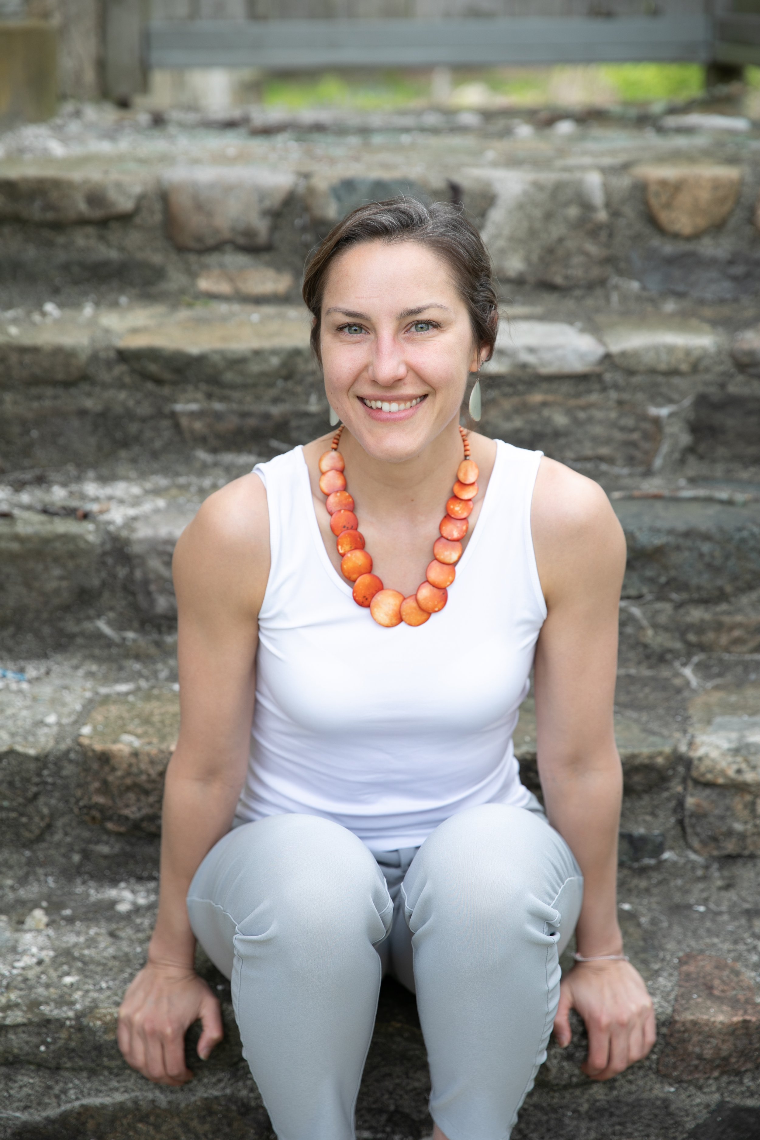 Trisha Mitchner Certified High Performance Coach™ A woman with short brown hair, wearing a white sleeveless top and light gray pants, sitting on stone steps outdoors, smiling at the camera, accessorized with large earrings and a beaded necklace.