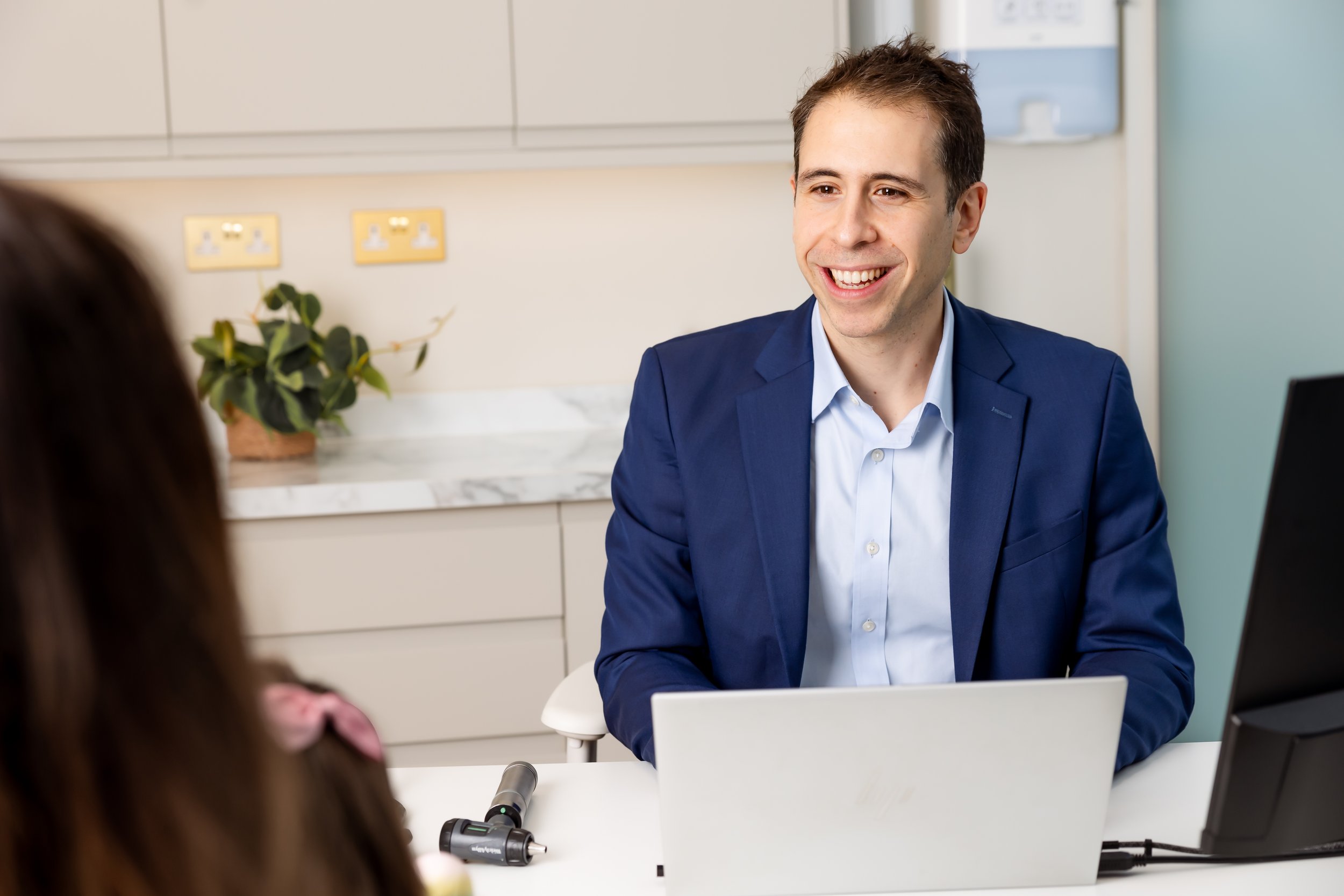 A man in a blue suit smiling while speaking to a woman in an office setting. The woman is partially visible in the foreground, and there is a potted plant and electrical outlets in the background.