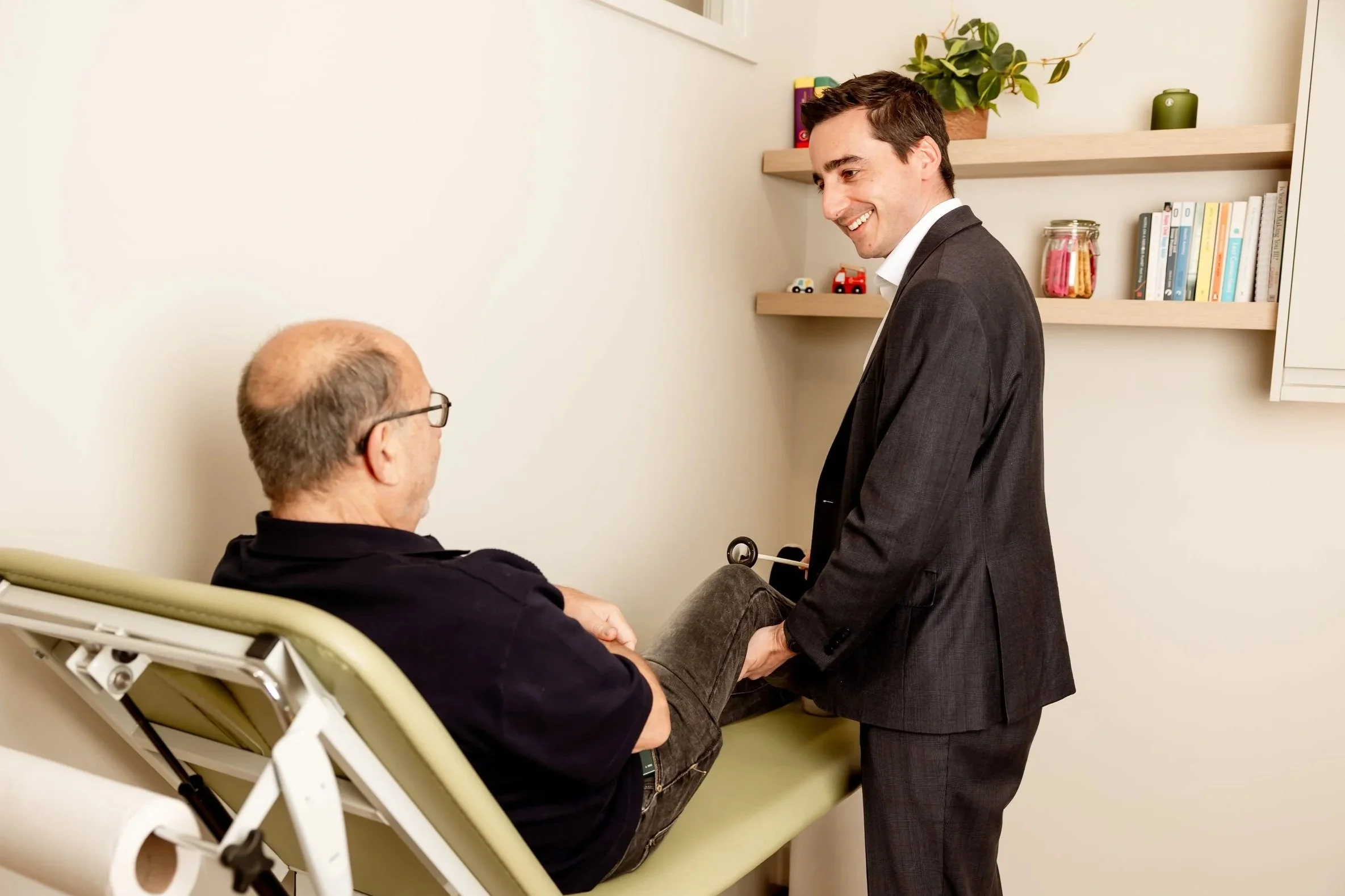 A young man in a gray suit is smiling and holding hands with an older man in a hospital bed. The older man is wearing glasses and a dark shirt, sitting in a hospital bed. There are shelves with books, a potted plant, and decorative items in the background.