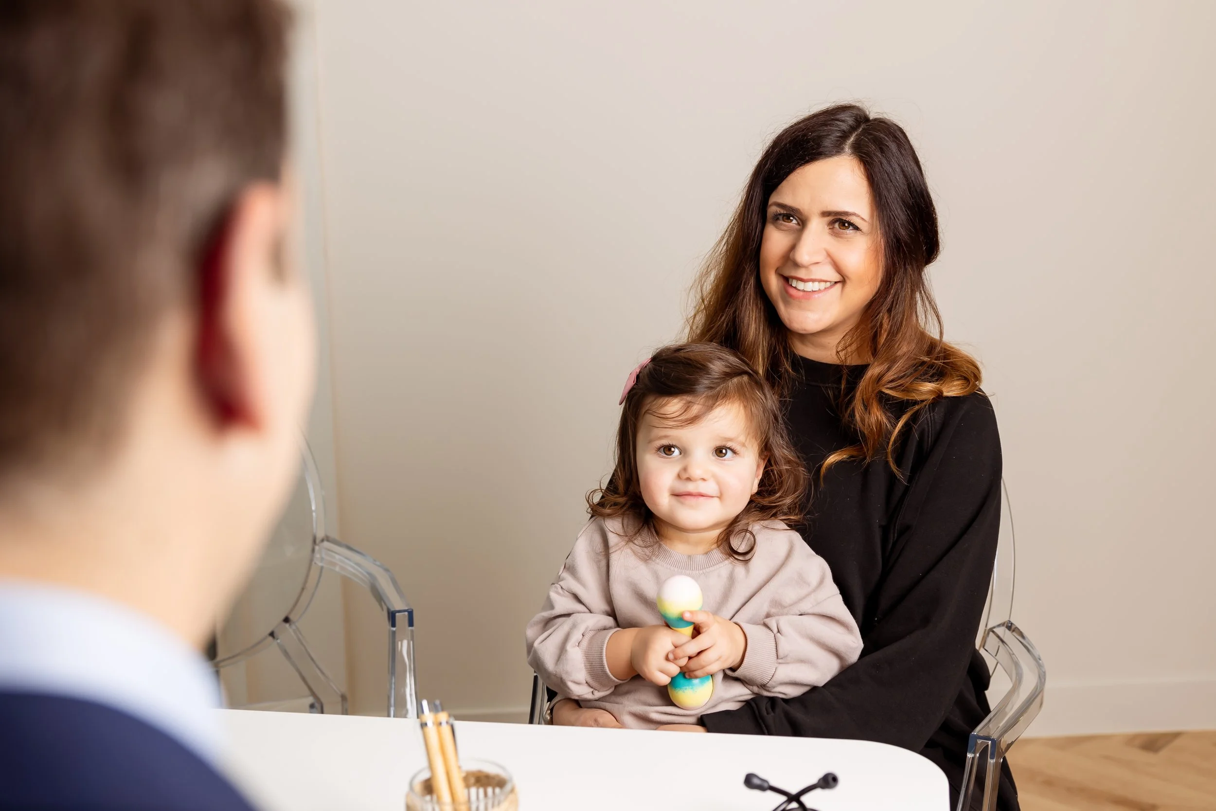 A woman with long brown hair and a child with curly hair and a pink bow, sitting at a table during an interview or consultation, smiling at a person across from them out of view.
