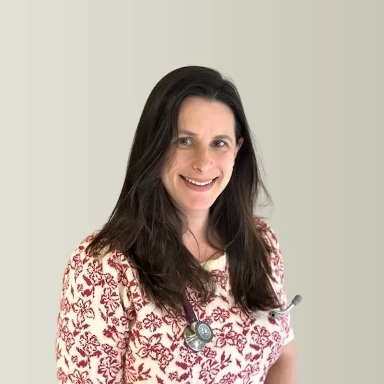 A woman with long dark hair wearing a patterned blouse and a stethoscope around her neck, smiling at the camera against a plain background.