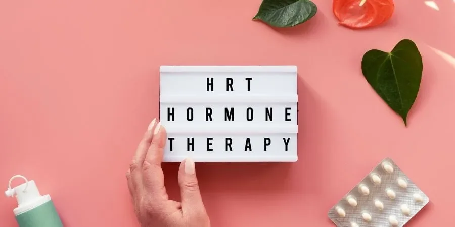 A lightbox sign with the words 'HRT', 'HORMONE', and 'THERAPY' on a pink background, surrounded by green leaves, a pink flower, a blister pack of white pills, and a small green bottle.