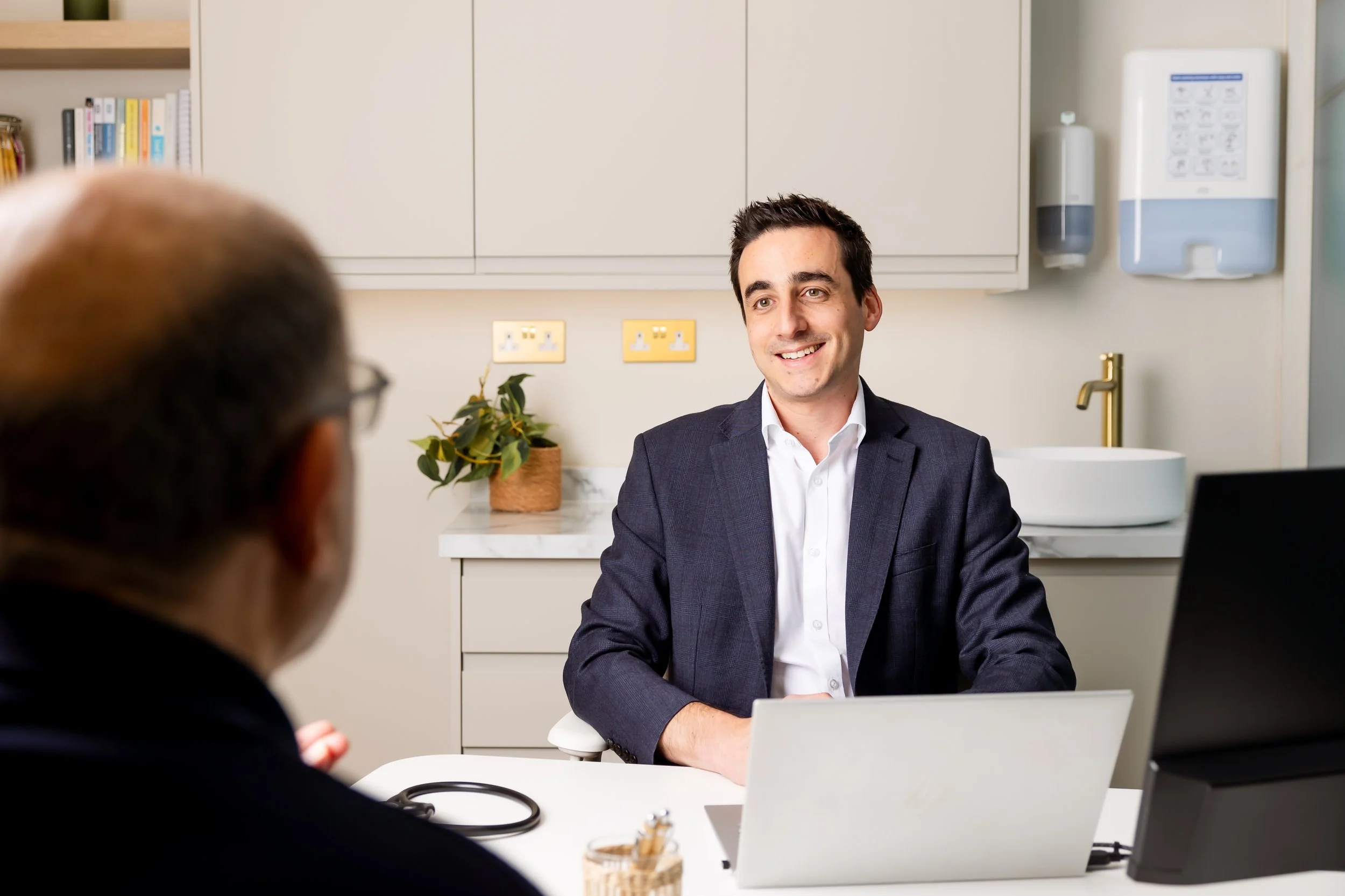 A man in a suit is sitting at a desk, smiling and talking to another person whose back is visible. The setting appears to be an office or medical clinic with cabinets, a plant, and medical equipment on the wall in the background.