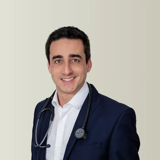 A smiling male doctor with dark hair, wearing a white shirt and a navy blue blazer, with a stethoscope around his neck, standing against a neutral background.