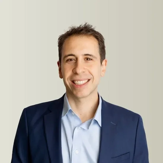 A smiling young man with short brown hair, wearing a light blue dress shirt and a dark blue suit jacket, standing against a neutral background.