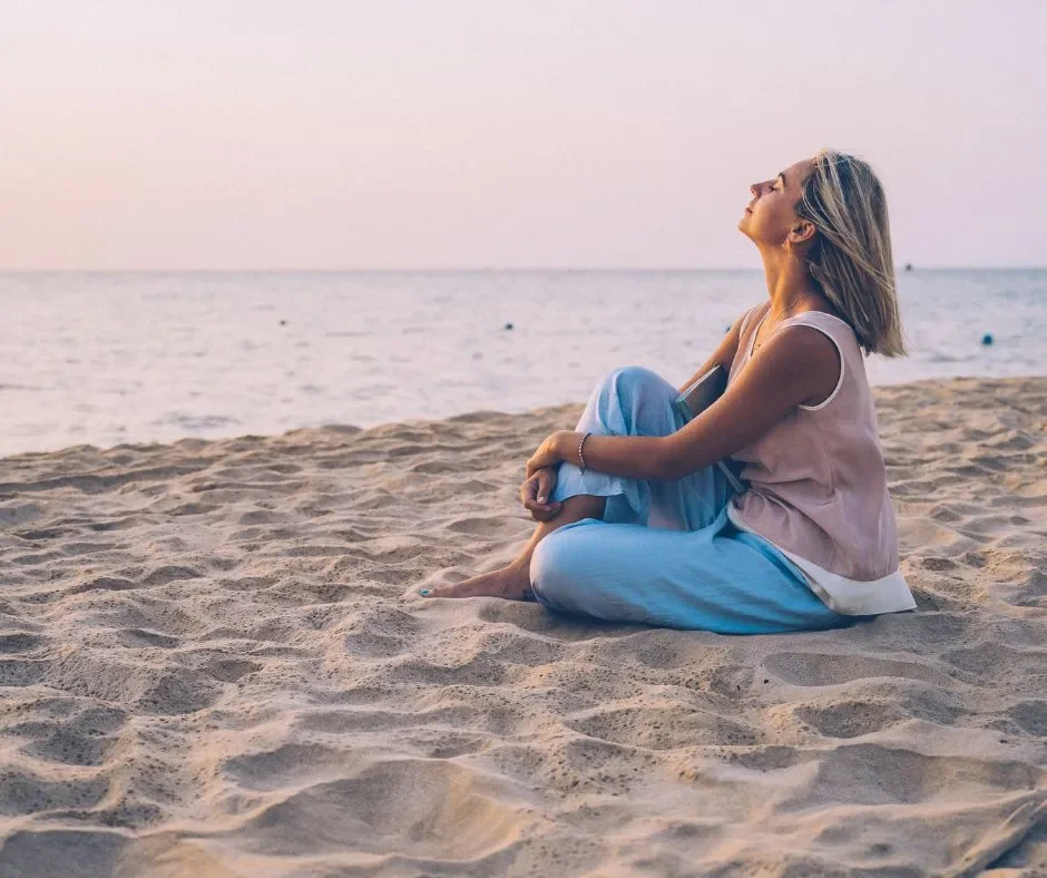 A woman sitting on the sandy beach near the ocean with her eyes closed and face tilted up, enjoying the sunlight.