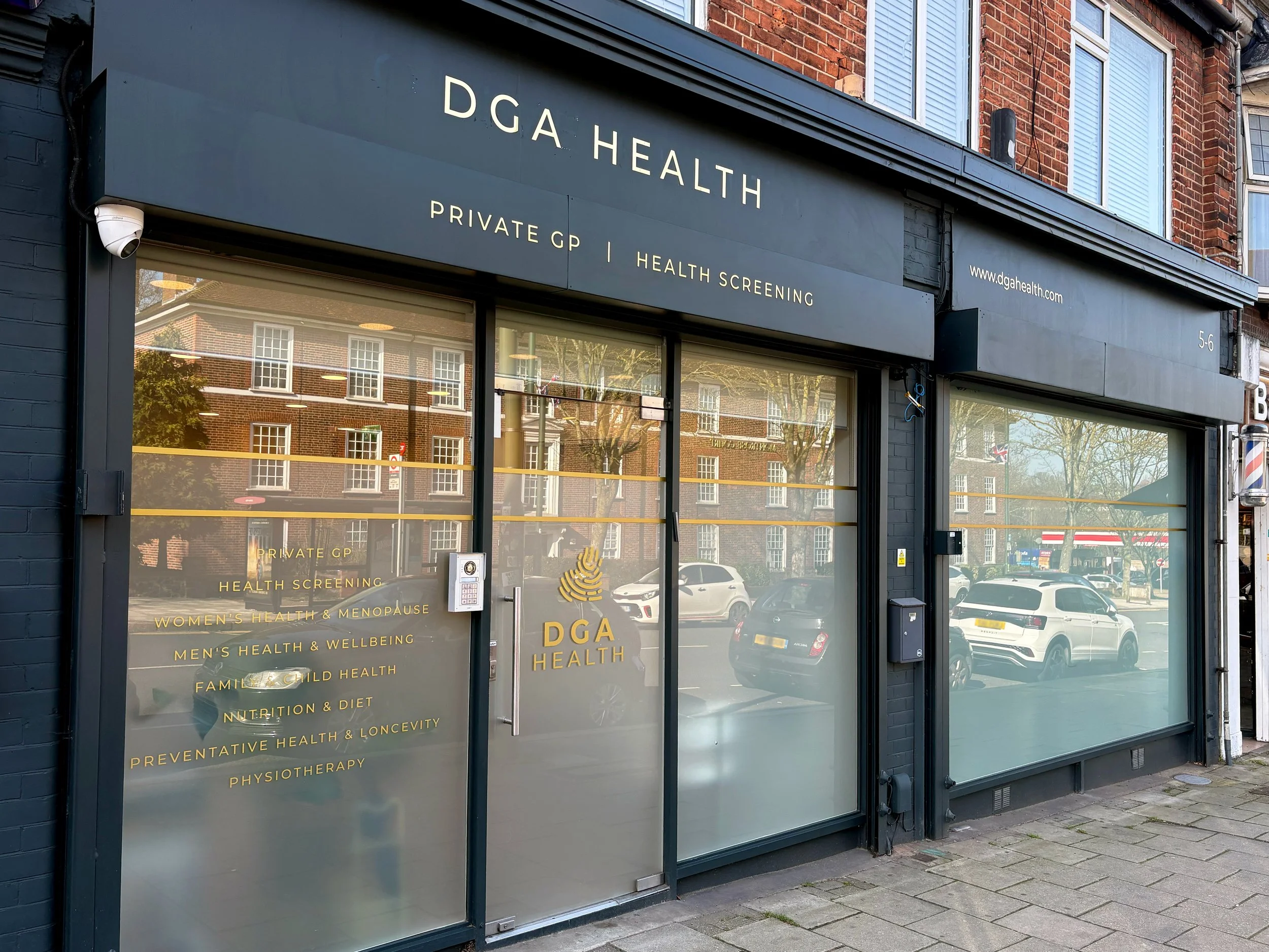 Exterior of DGA Health clinic with a dark gray storefront, large windows, and a sign displaying services like private GP, health screening, women's health, men's health, family health, nutrition, preventative health, physiotherapy, and osteopathy. The building has a red brick upper level and a security camera near the top left corner.