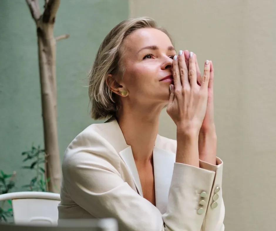 A woman with short, gray hair and light skin, wearing a white blazer, is sitting with her eyes closed and hands pressed together near her face, in a peaceful outdoor setting with a light green wall and a small tree in the background.