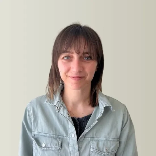 A woman with shoulder-length brown hair and bangs, wearing a light denim jacket over a dark top, smiling at the camera against a plain beige background.