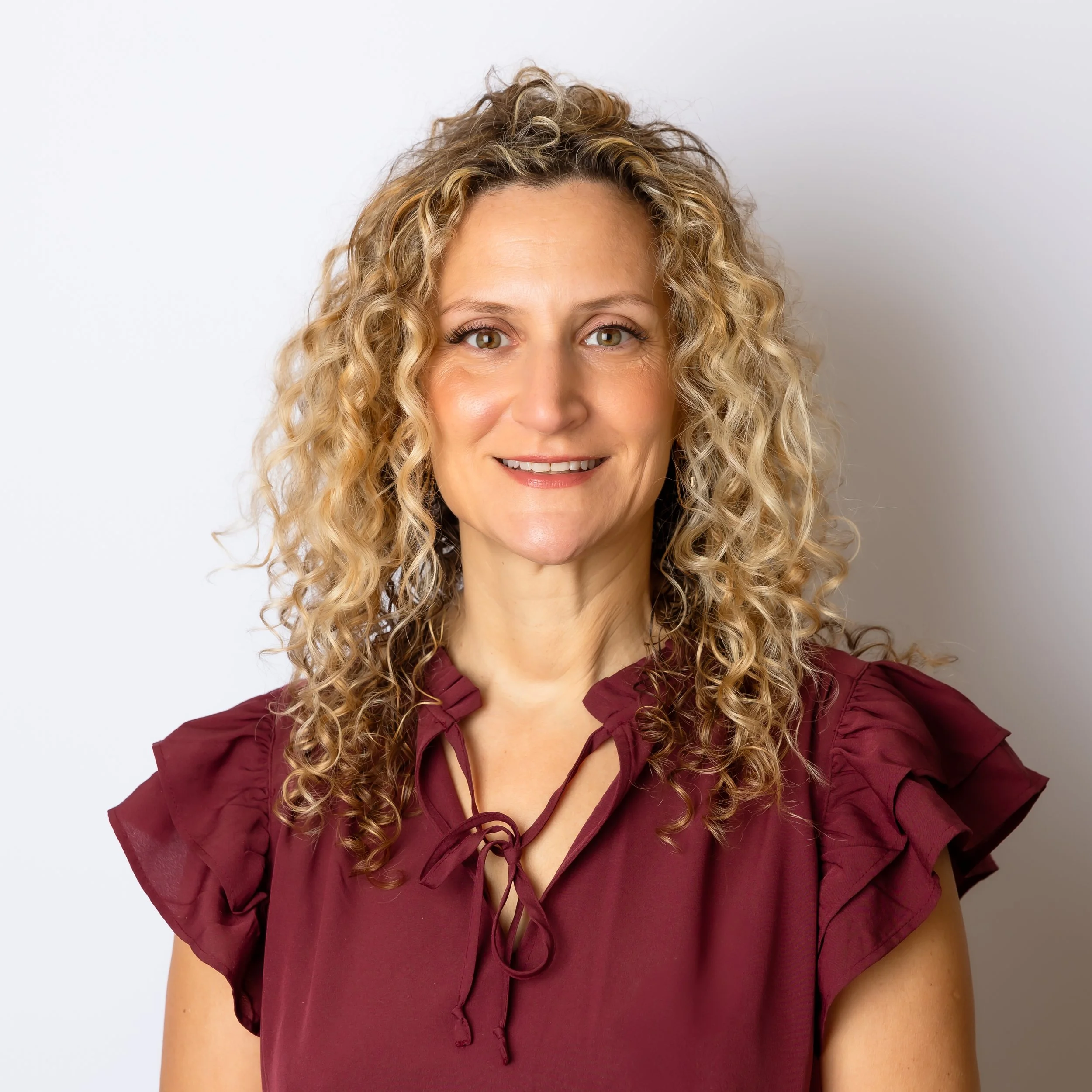 Close-up of a woman with curly blonde hair, wearing a maroon blouse with lace-up detail, smiling, against a plain white background.
