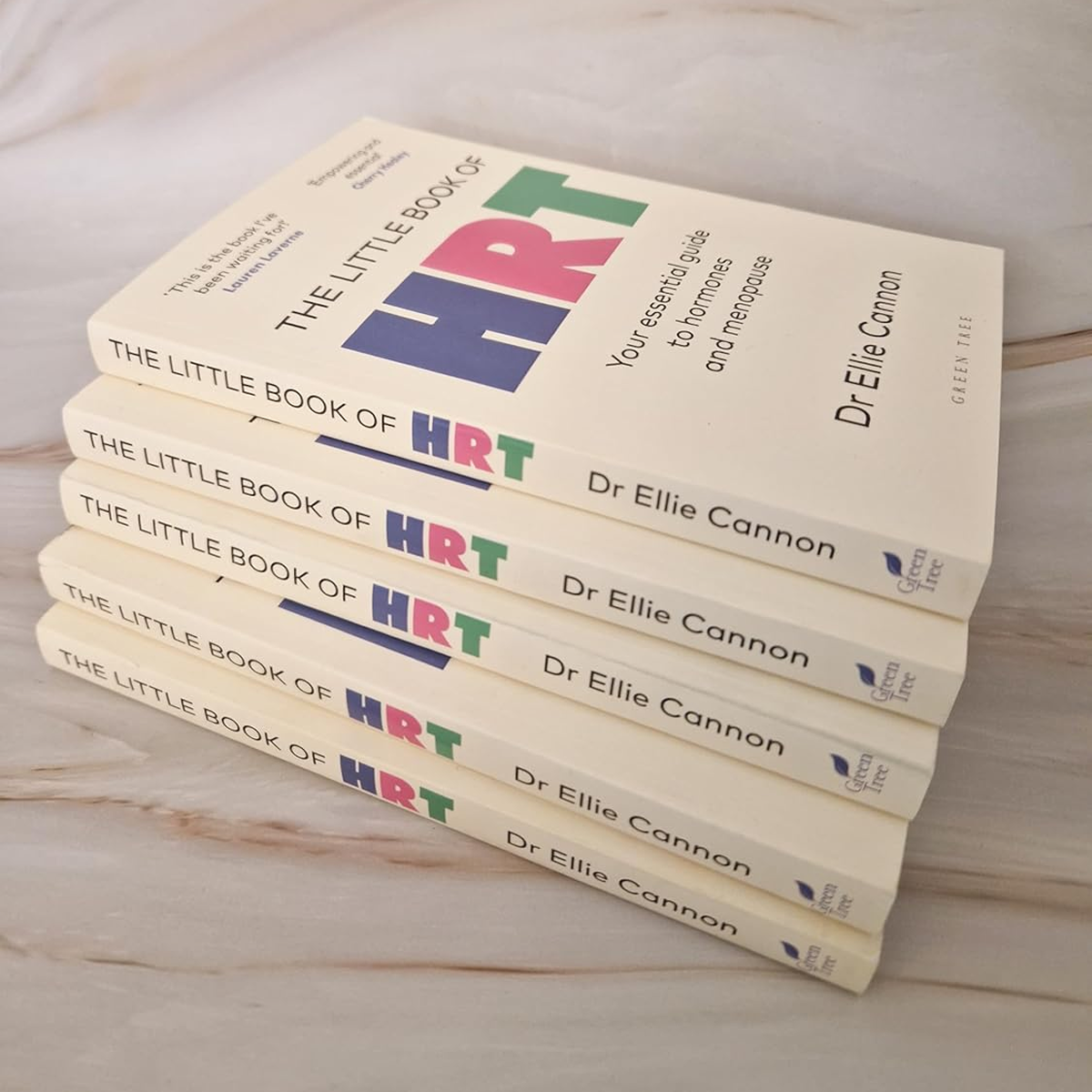 Stack of five copies of the book titled 'The Little Book of HRT' by Dr. Ellie Cannon, with a white cover and colorful text, on a light wooden surface.