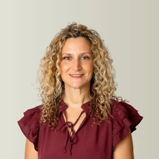 A woman with curly blonde hair wearing a burgundy top with ruffled sleeves, smiling at the camera against a neutral background.