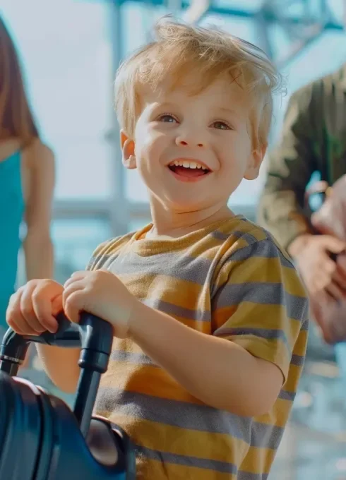 A young boy with blonde hair smiling and holding onto a stroller indoors, with blurred people and large windows in the background.