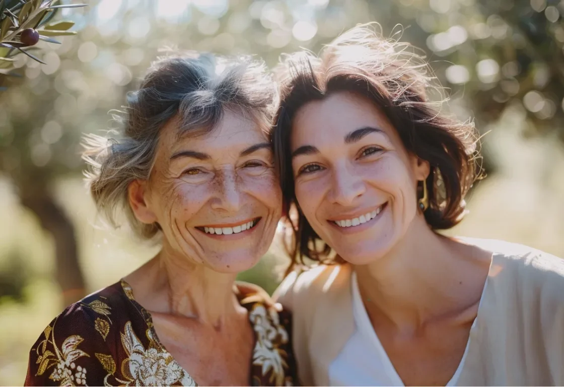 Two women smiling outdoors in a sunny setting with trees in the background.