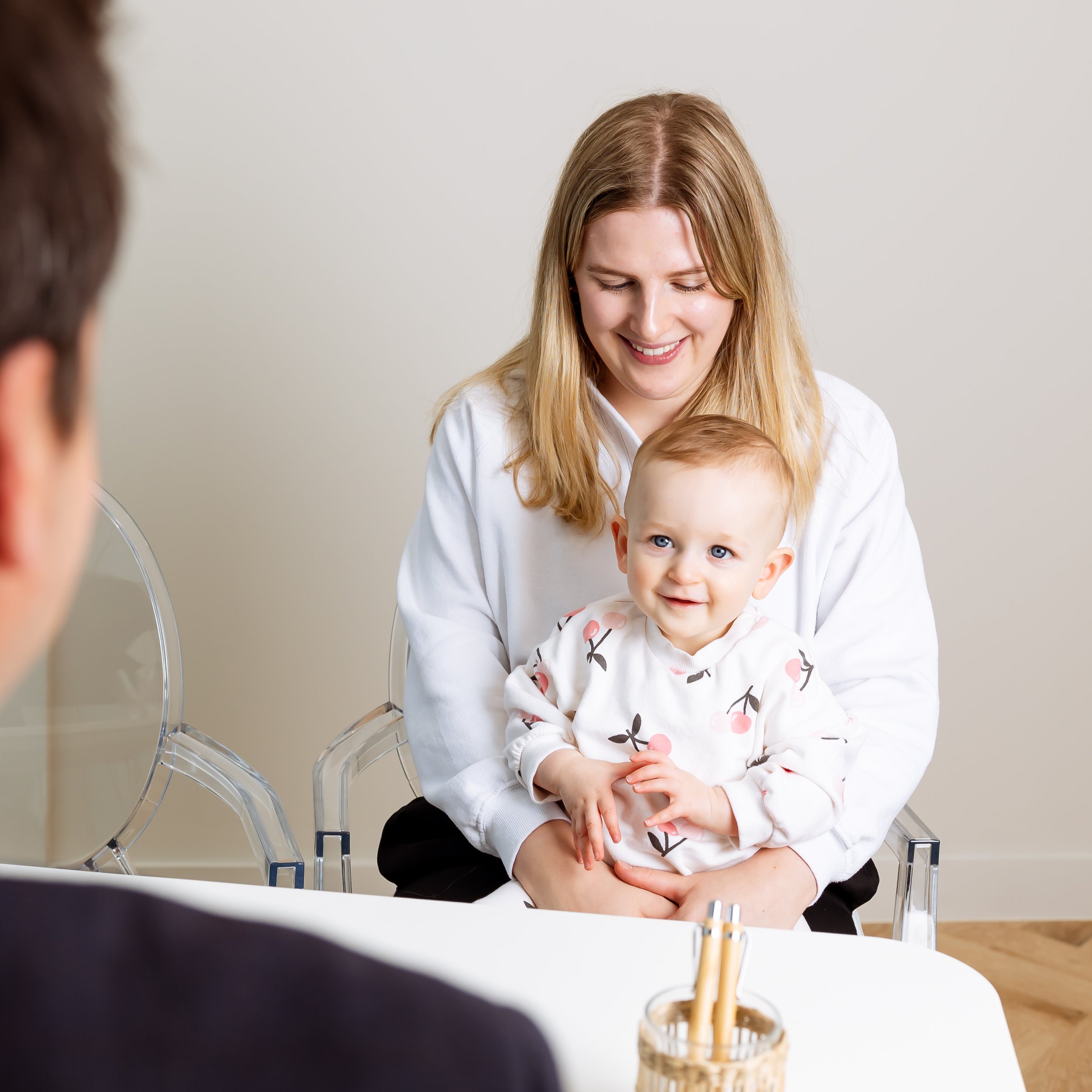 A woman with long blonde hair smiling while sitting across from a young child with blue eyes and light brown hair, in a bright room with neutral-colored walls. There are transparent chairs and a table with a glass filled with straw and a beverage, suggesting a casual or professional setting.