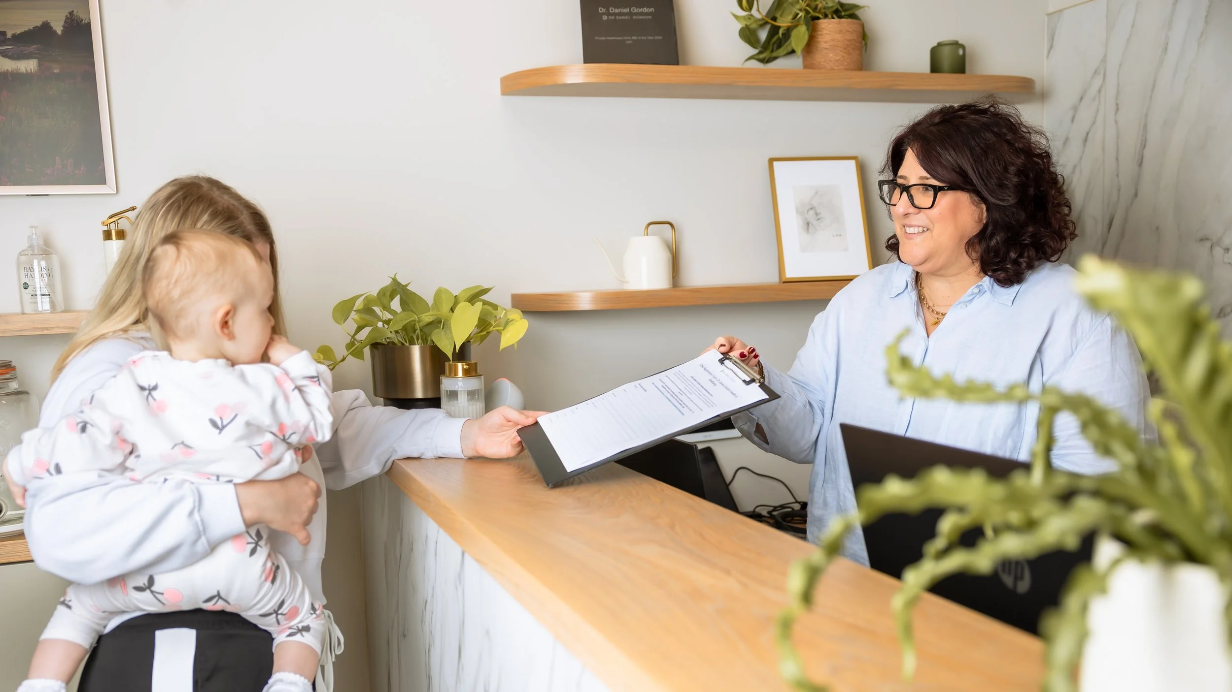 A young girl is sitting on a woman's lap at a reception desk, handing over papers to a receptionist. The woman is smiling and wearing glasses and a blue shirt. The background has shelves with decorative items and framed pictures, and there are plants on the counter.