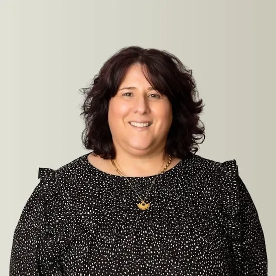 A woman with shoulder-length dark curly hair, wearing a black and white polka dot blouse and a gold necklace with a small pendant, smiling in front of a light-colored background.
