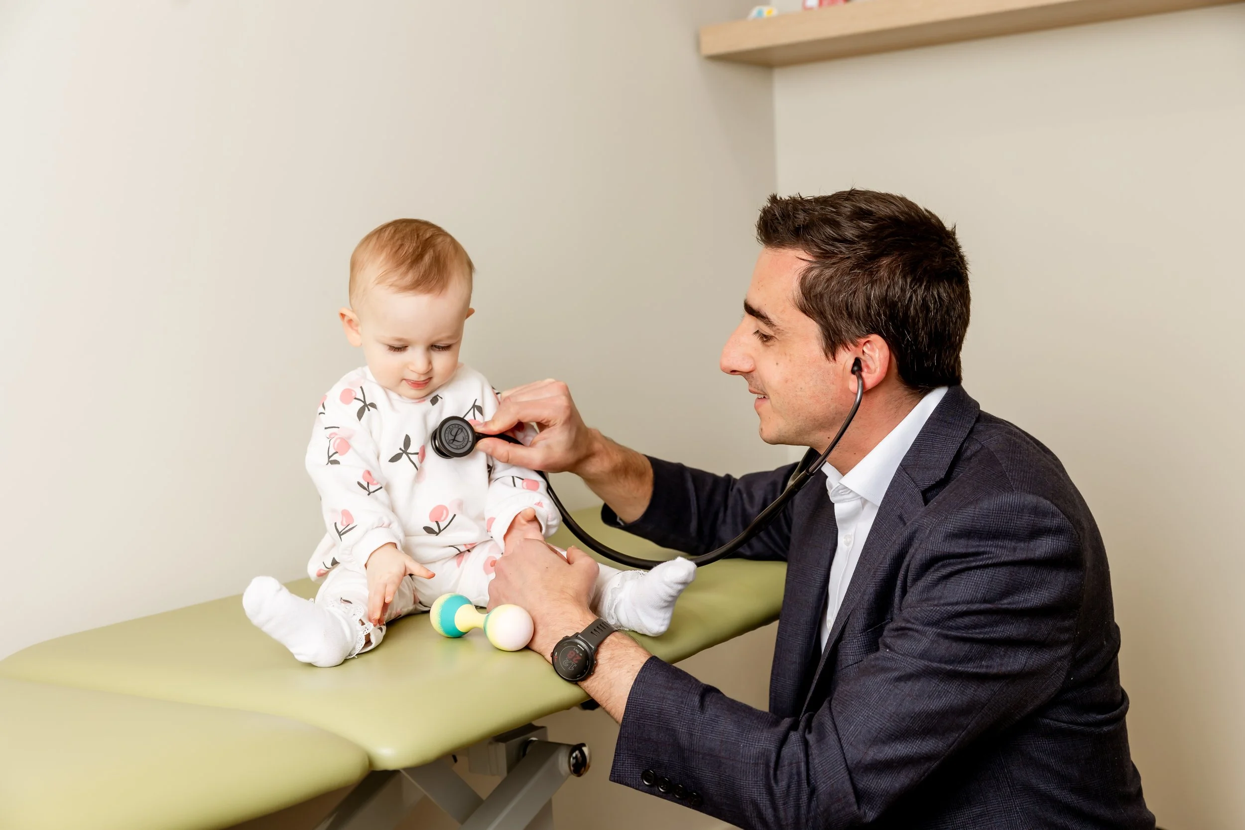 A young girl sitting on an examination table while a male doctor with a stethoscope checks her heartbeat. The girl is wearing pajamas with a flower pattern, and the doctor is smiling. The background is a plain wall with a shelf.