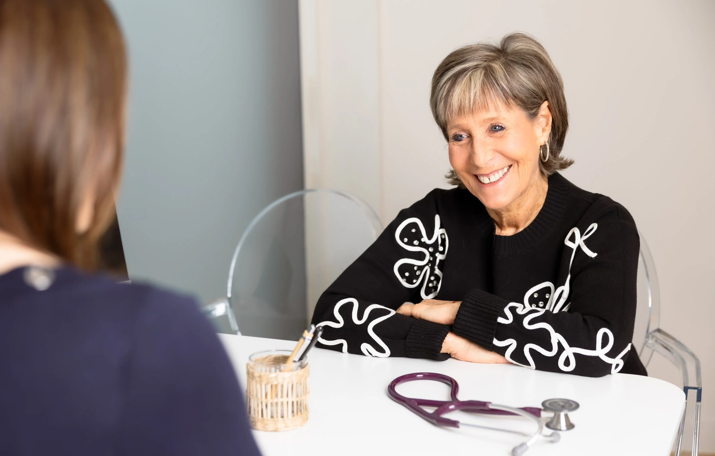 A smiling older woman with short gray hair and blue eyes, wearing a black sweater with white butterfly designs, sitting at a white table with a stethoscope and a small wicker basket, talking to a person with reddish-brown hair and a dark blazer.