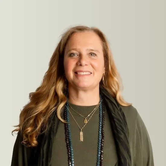 A woman with long wavy blonde hair smiling, wearing multiple necklaces and a dark jacket over a green top, standing against a light gray background.
