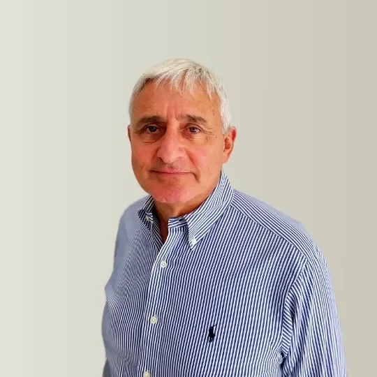A middle-aged man with gray hair and light skin, wearing a blue and white striped button-up shirt, standing against a plain light background.