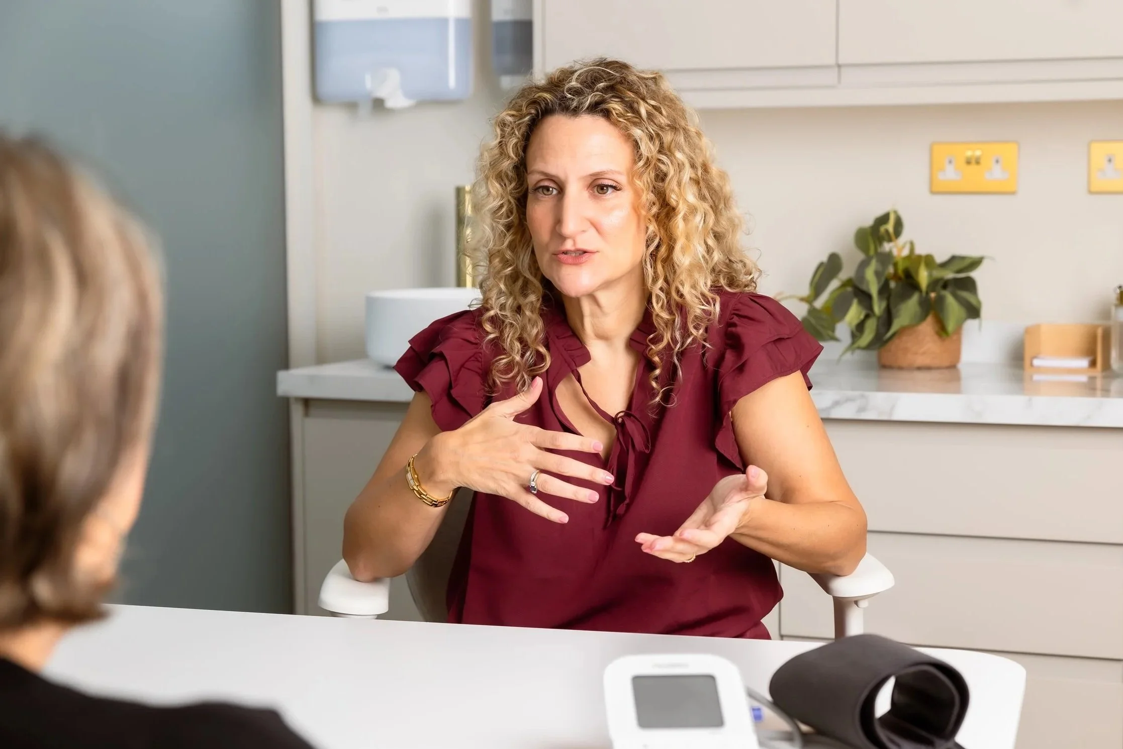 A woman with curly blonde hair, wearing a maroon blouse, is speaking and gesturing with her right hand during a conversation in a medical setting, with another person partially visible in the foreground. The background includes a white countertop, a plant in a woven pot, and medical electrical outlets.
