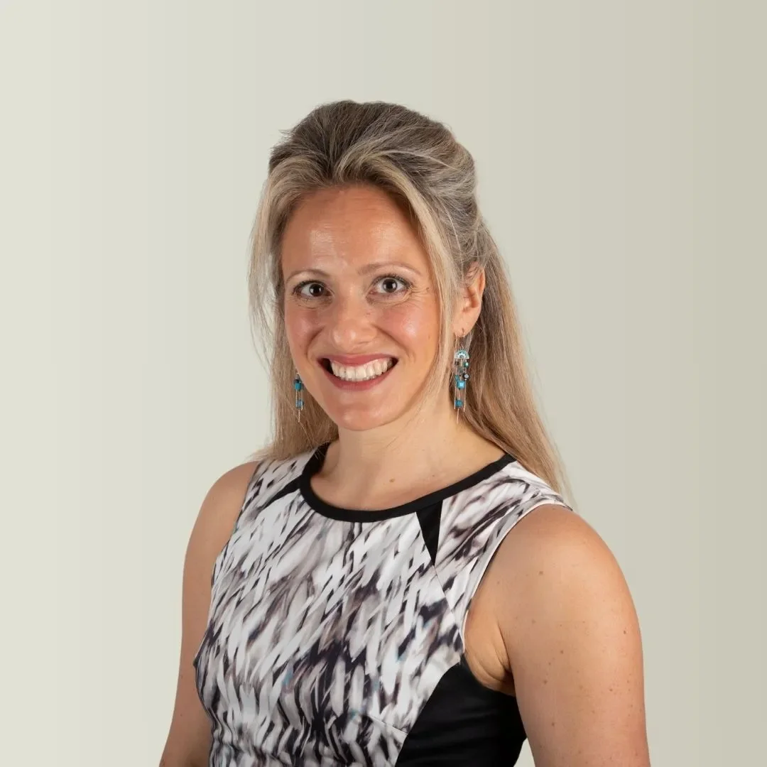 A woman with long blonde hair wearing a sleeveless black and white patterned top and blue earrings, smiling at the camera.