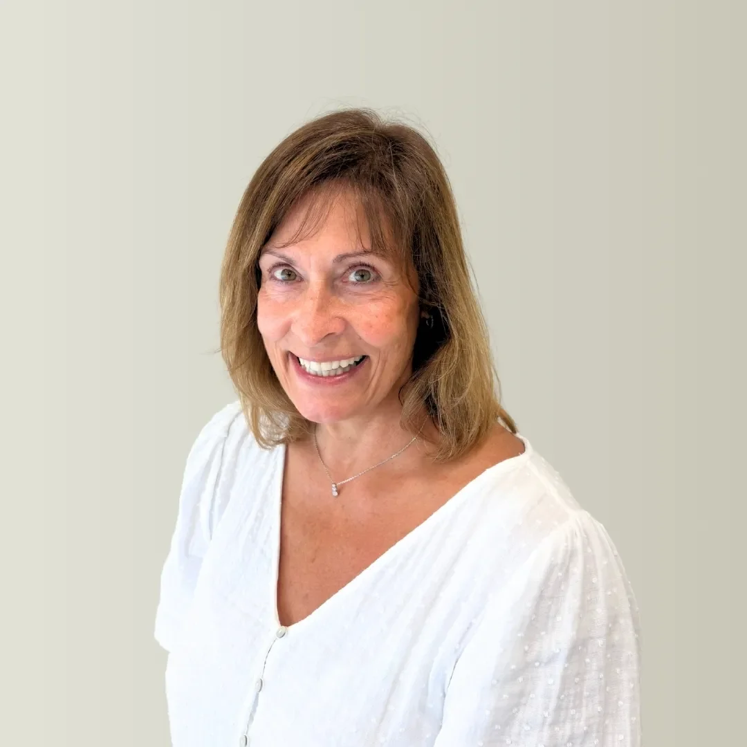 A smiling middle-aged woman with shoulder-length brown hair wearing a white blouse and a delicate necklace, standing against a light background.