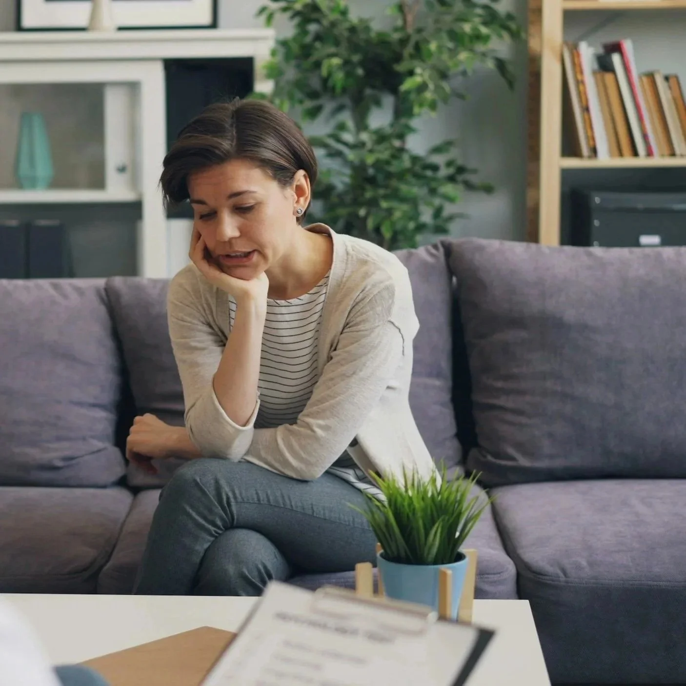 A women wearing a striped shirt and jeans seated on a sofa and looking down. She is talking.