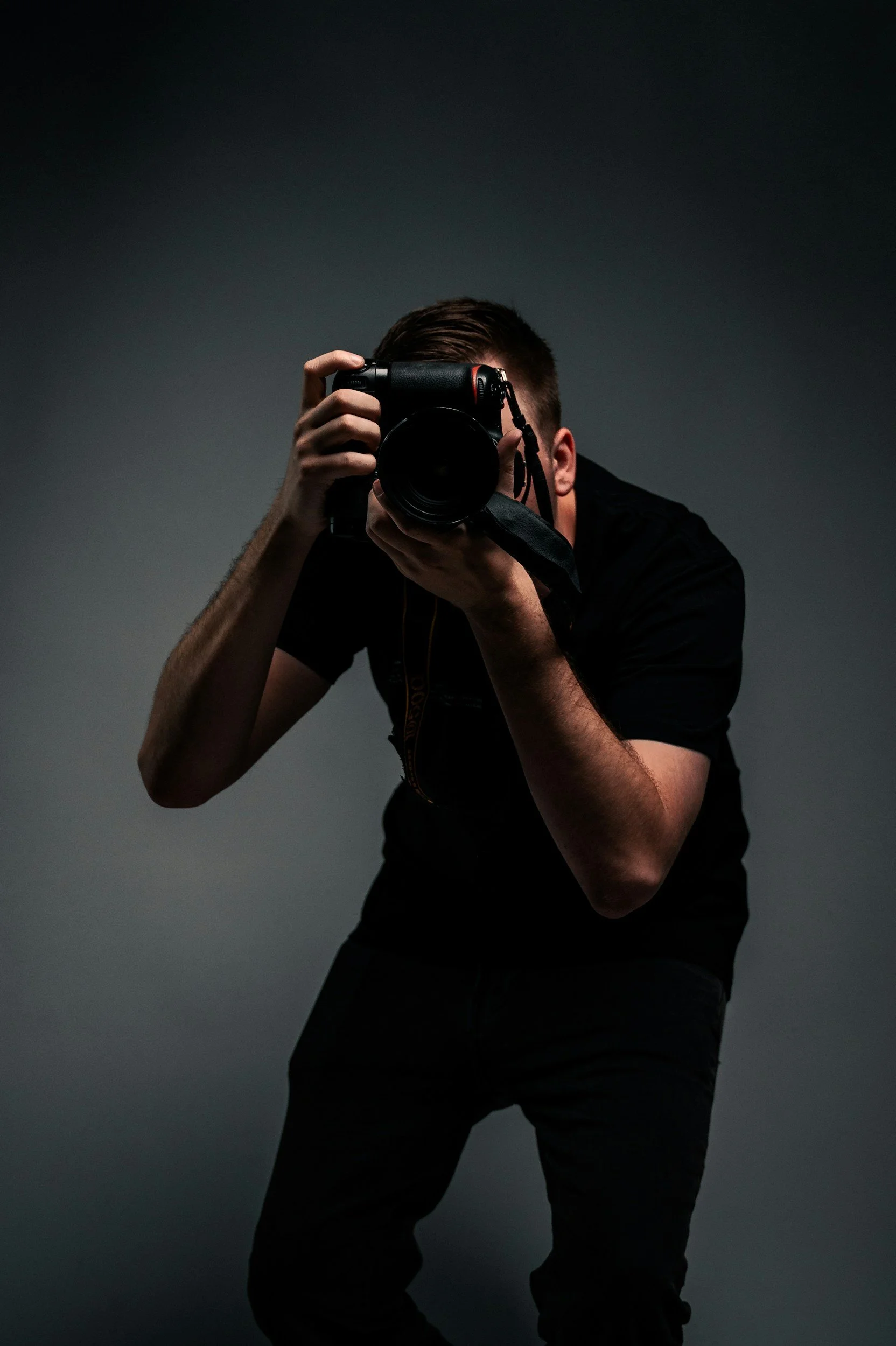 Photographer, holding a professional camera with a large lens, aiming it toward the viewer, against a plain dark background.