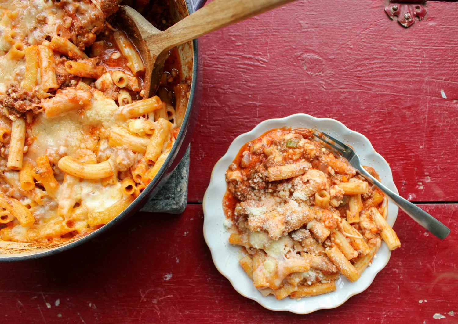 Baked pasta casserole dish with cheese, served next to a plate of the same pasta topped with grated cheese and a fork, on a red wooden table.