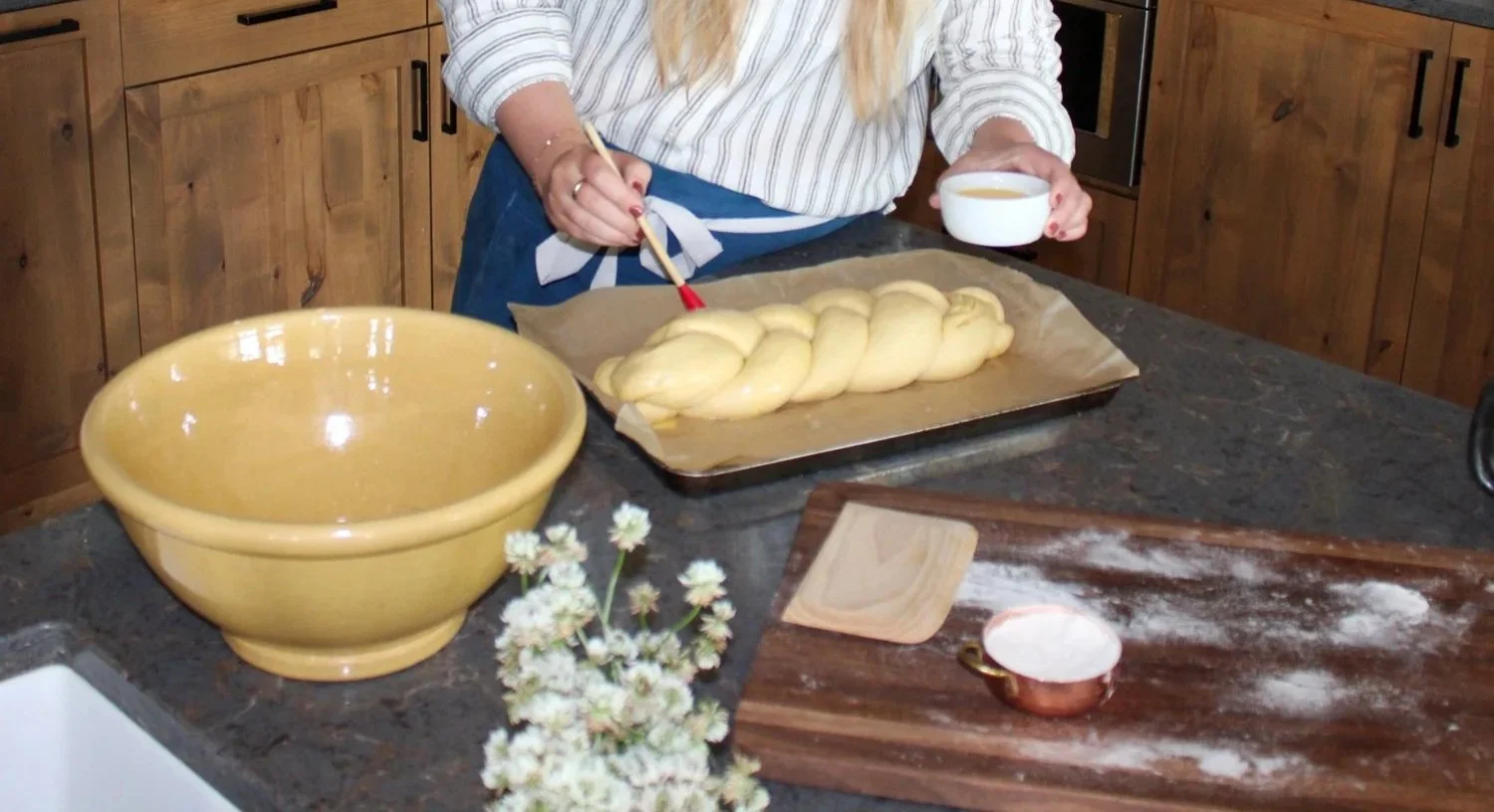 A person is twisting bread dough on a baking sheet in a kitchen, holding a bowl, with flowers, a flour-dusted cutting board, and a large yellow mixing bowl on the counter.