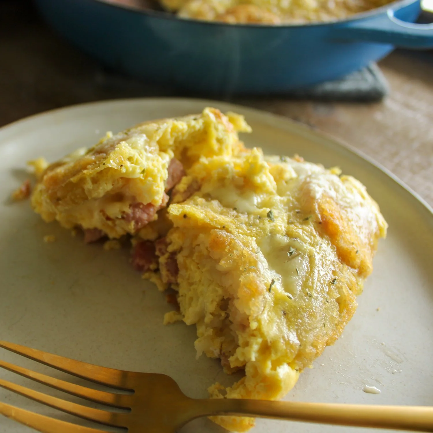 an egg casserole sitting on a cream plate with a gold fork, sitting in front of a blue pan