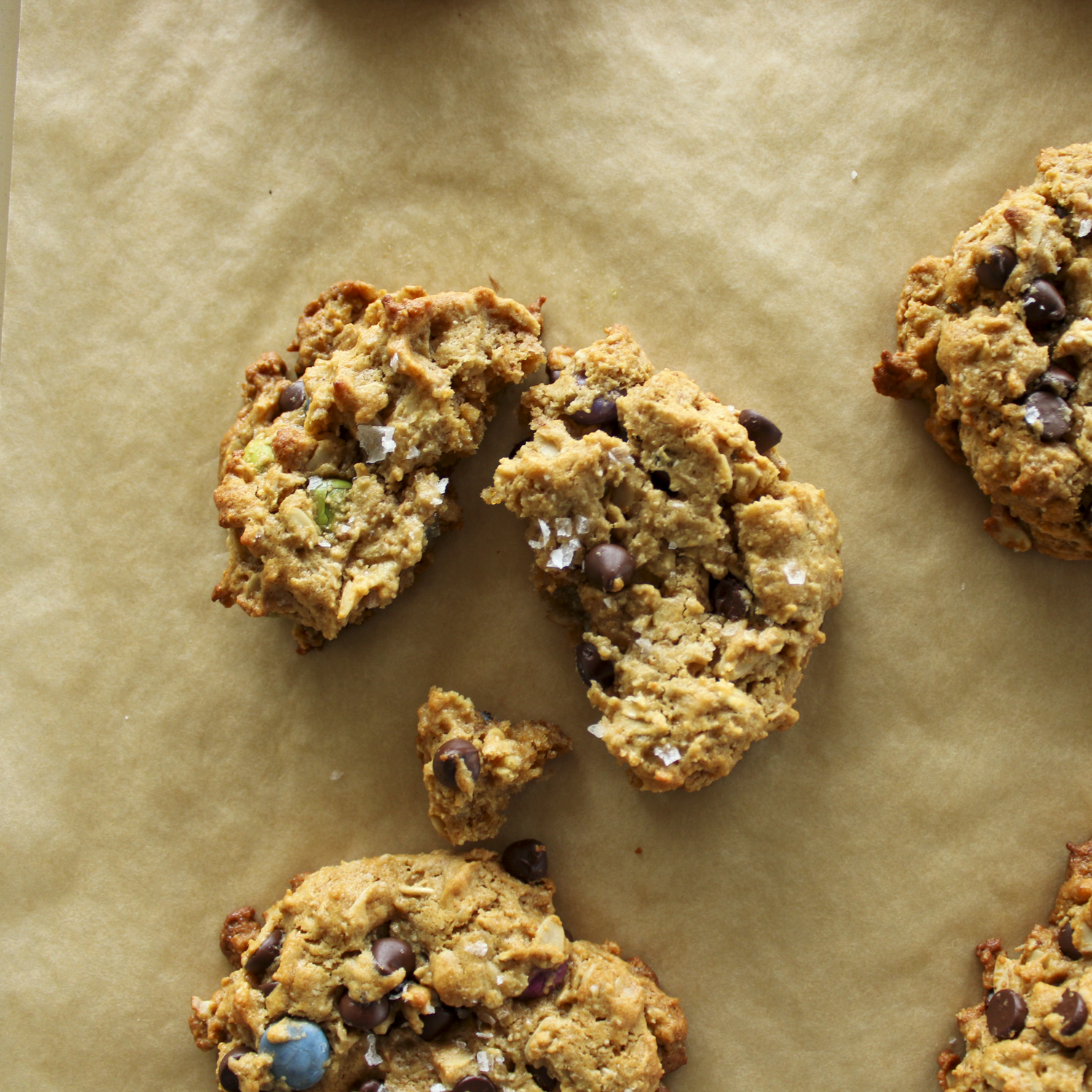 a monster cookie topped with sea salt, broken in half, sitting on parchment paper with other cookies