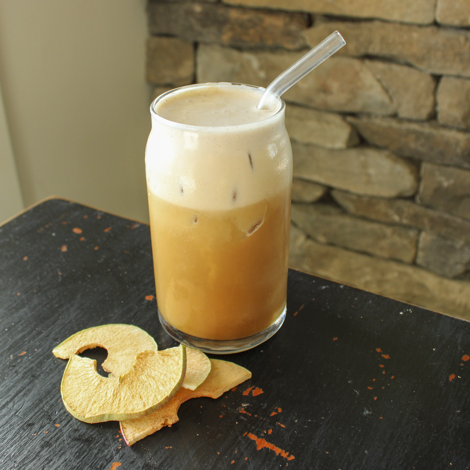 a cold brew coffee in a glass cup with a glass straw, sitting on a black chipped table with dried apples next to it
