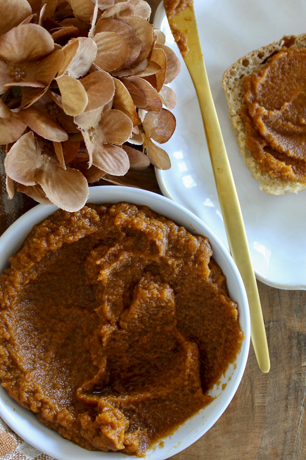a white bowl of pumpkin butter sitting on a wood board with a slice of toast slathered with pumpkin butter sitting on a cream plate with a gold knife