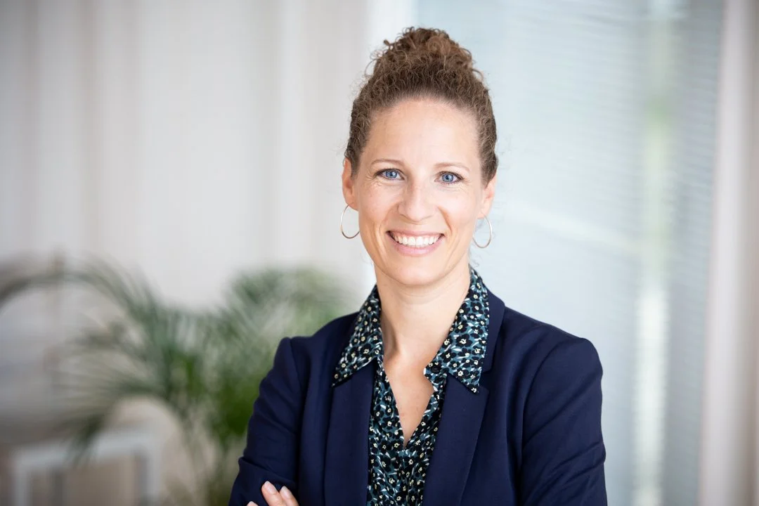 Eine lachende Frau mit lockigem Haar, die einen blauen Blazer trägt, vor einem Bürohintergrund.