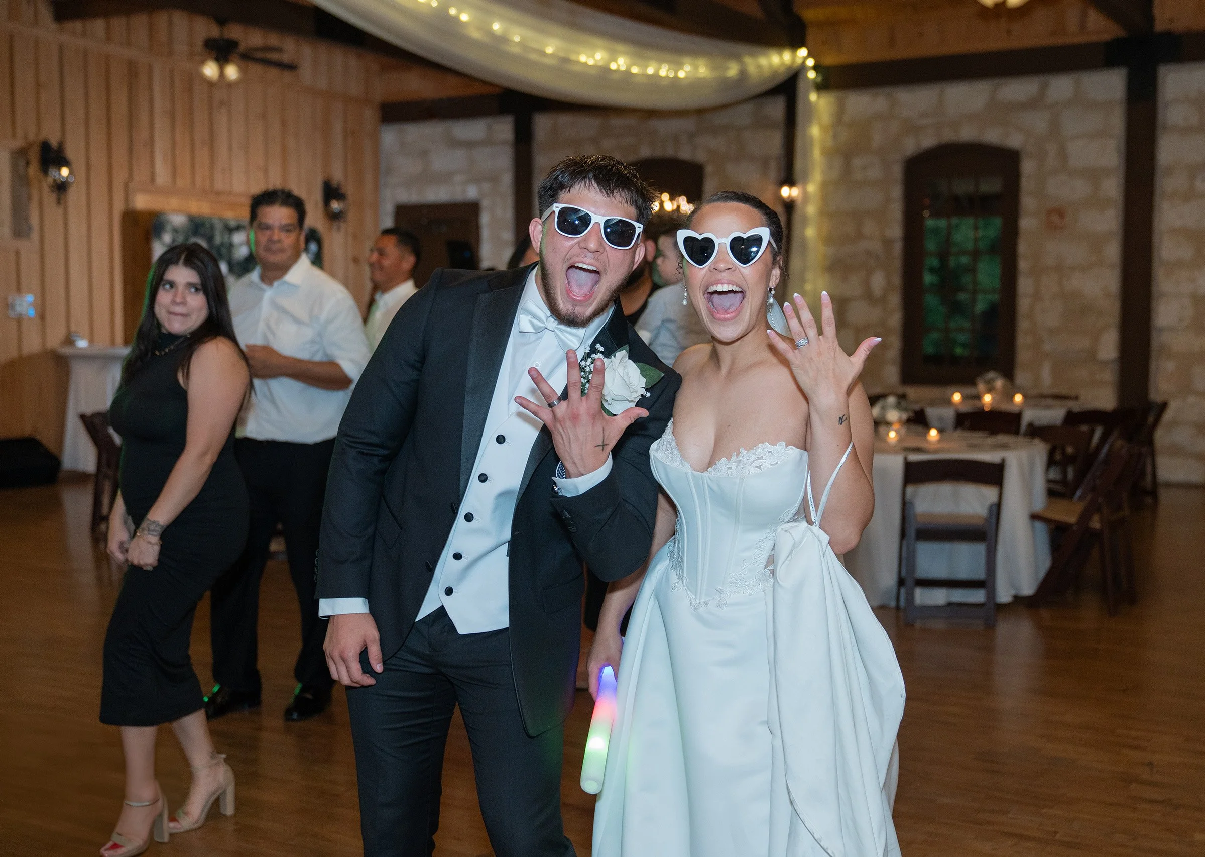 Wedding reception with a man and woman in tuxedo and wedding dress wearing sunglasses showing rings, celebrating indoors.