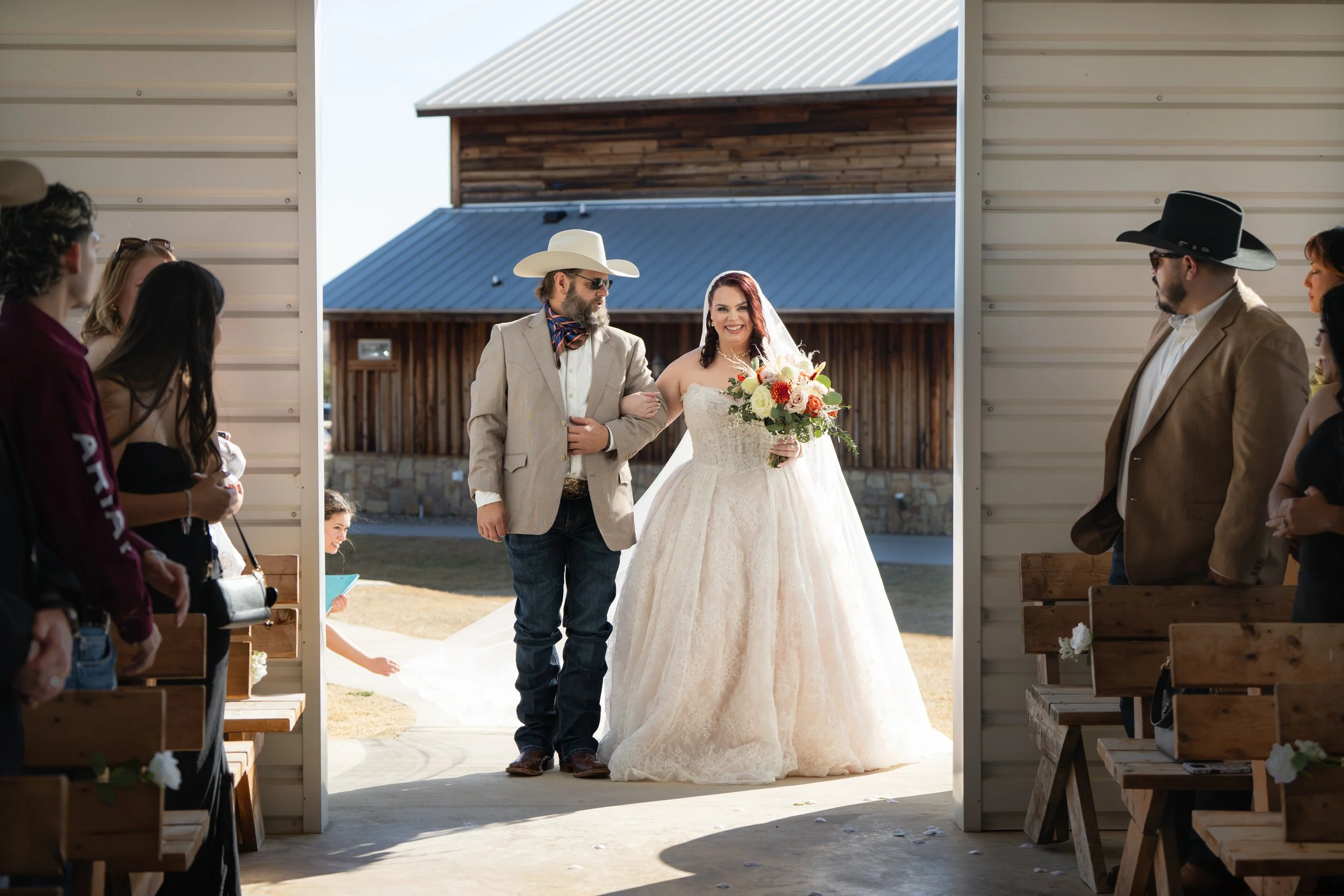 Bride walking down the aisle arm-in-arm with her father at an outdoor wedding ceremony, guests seated on either side, with rustic barn buildings in the background.