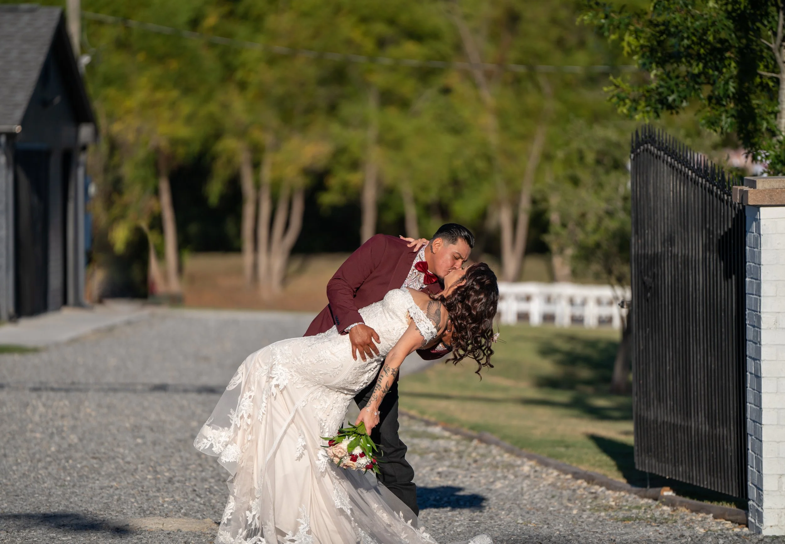 A groom and bride kiss outdoors during their wedding, with the groom in a maroon suit and the bride in a white lace wedding dress holding a bouquet of flowers.