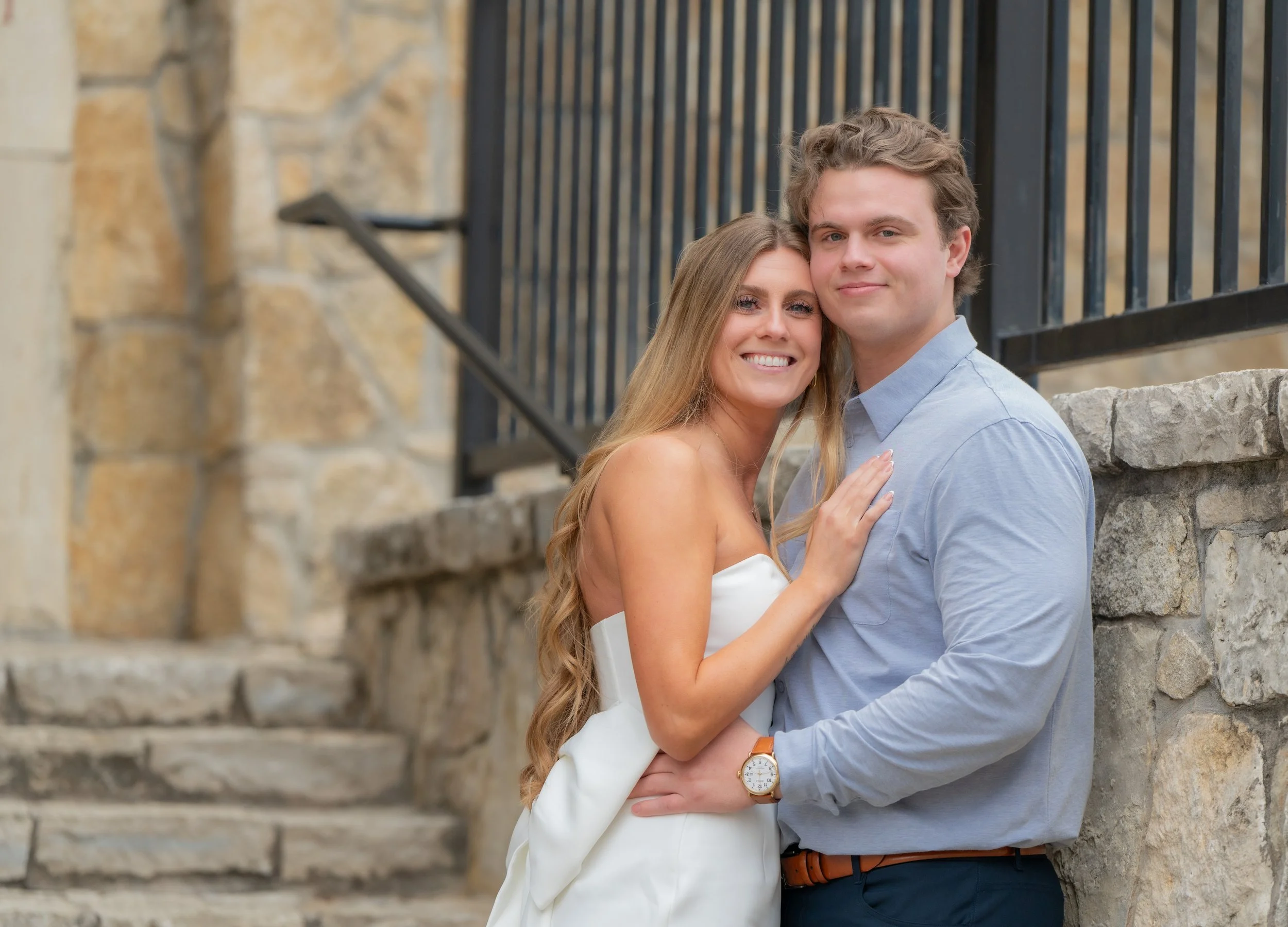 A smiling couple, a woman in a white dress and a man in a light blue shirt, standing close together outdoors near stone stairs and a black metal fence.