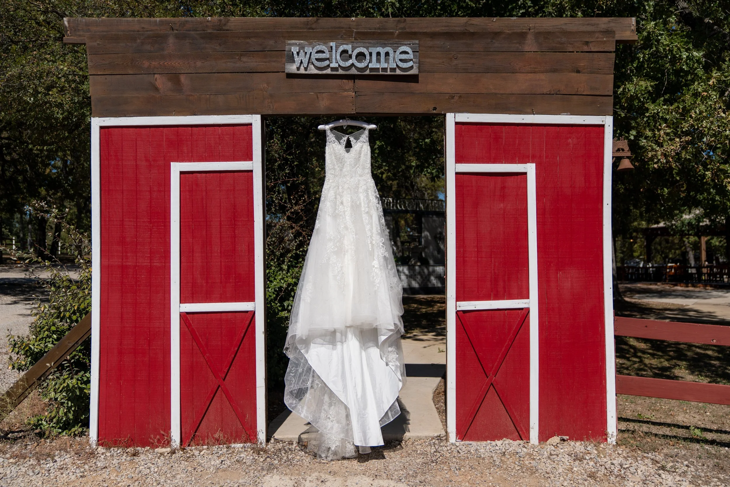 Wedding dress hanging on a hanger in front of a red and white barn-style backdrop with a sign that says 'welcome' on top.