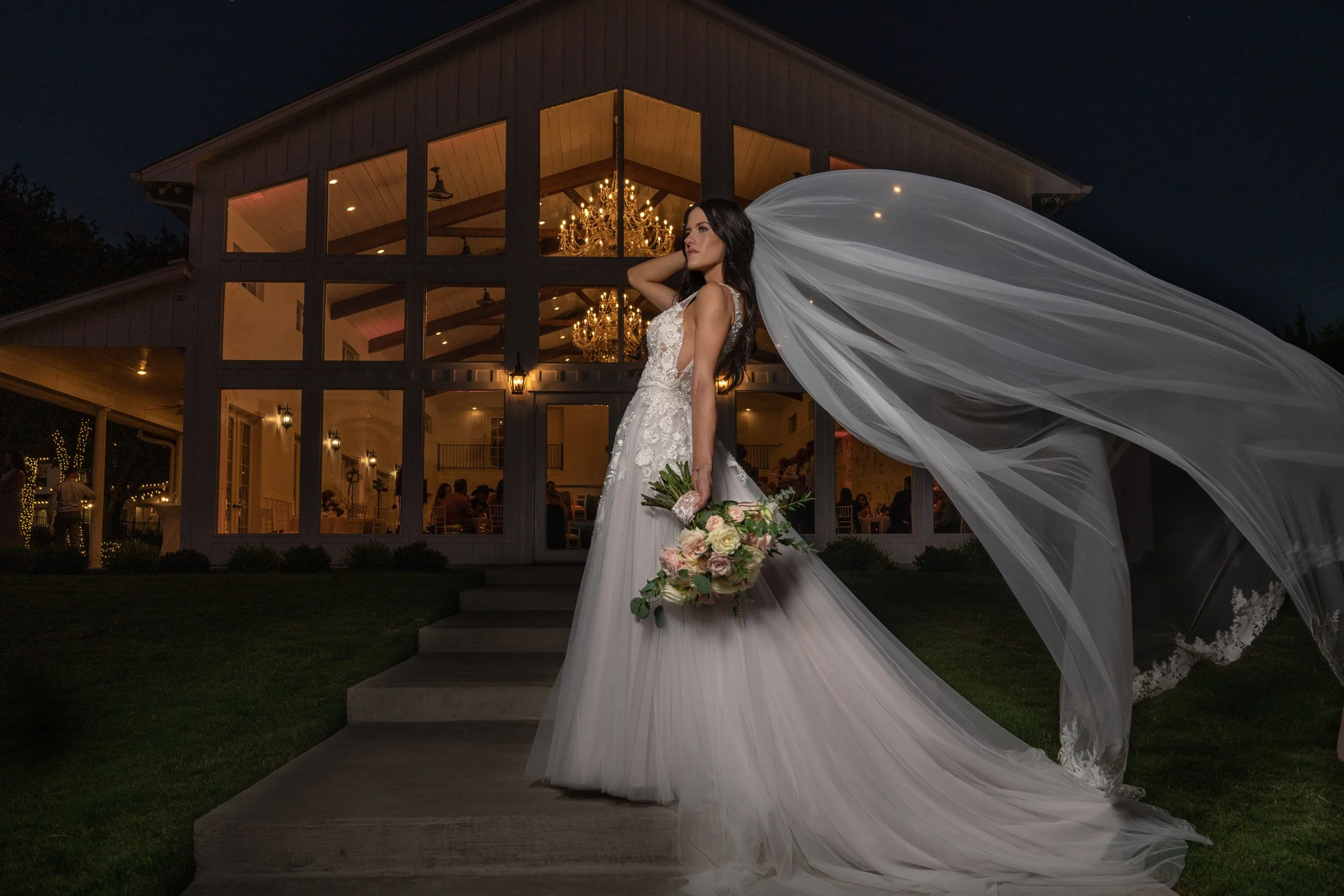 A bride in a wedding dress holding a bouquet of flowers, standing outside a lit-up building at night, with her veil flowing in the wind.