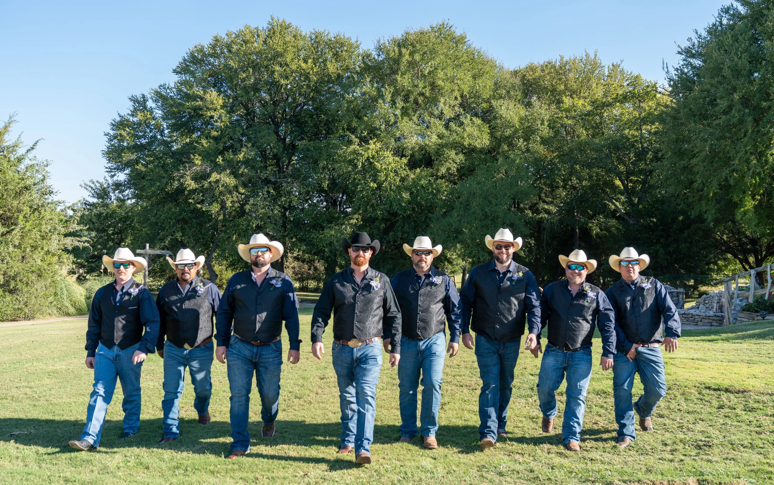 A group of men walking outdoors on a grassy area with trees in the background. They are wearing cowboy hats, sunglasses, and casual western-style clothing.