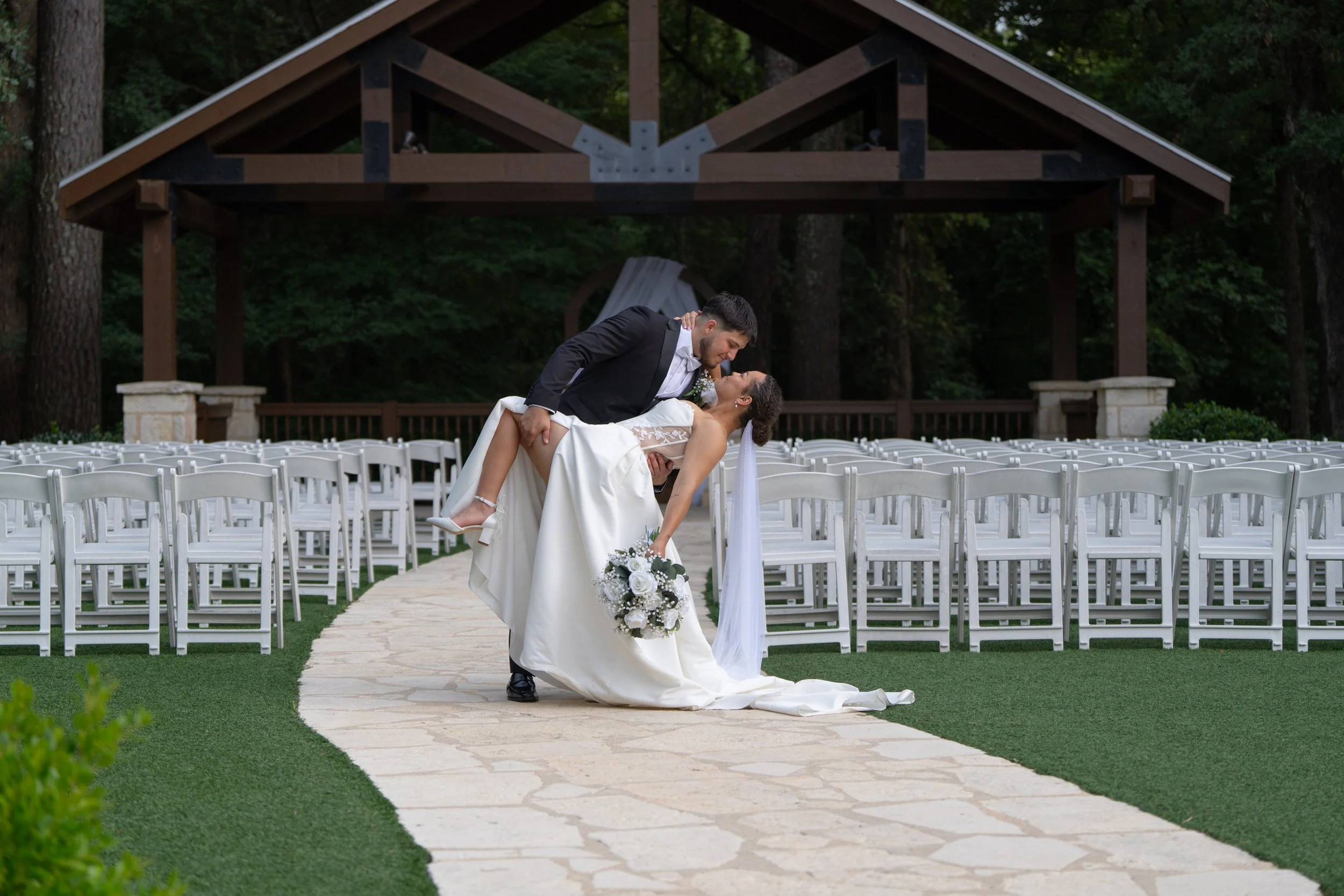 Bride and groom at an outdoor wedding venue, with the groom dipping the bride back in a dance pose, surrounded by white chairs on a stone pathway and a wooded backdrop.