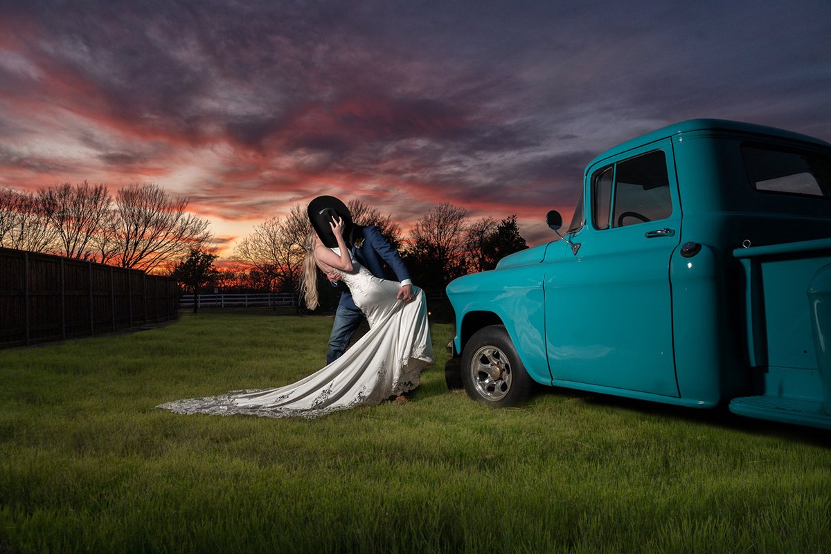 A person in a blue jacket and jeans kissing a bride in a white wedding dress on her wedding day in a grassy yard at sunset, with a vintage blue truck nearby.