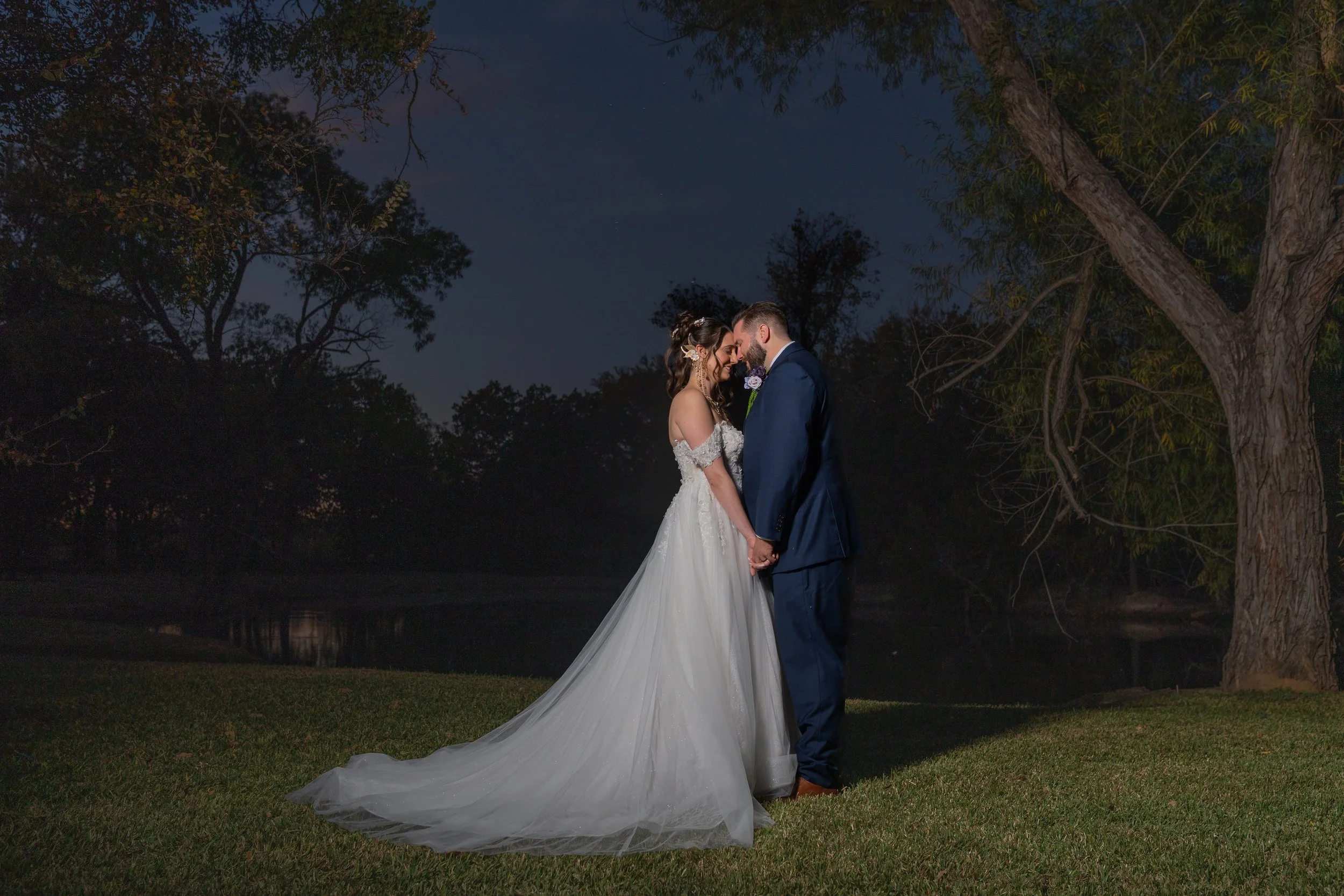 A bride and groom stand close together at night outdoors, touching foreheads and holding hands, with trees and a dark sky in the background.