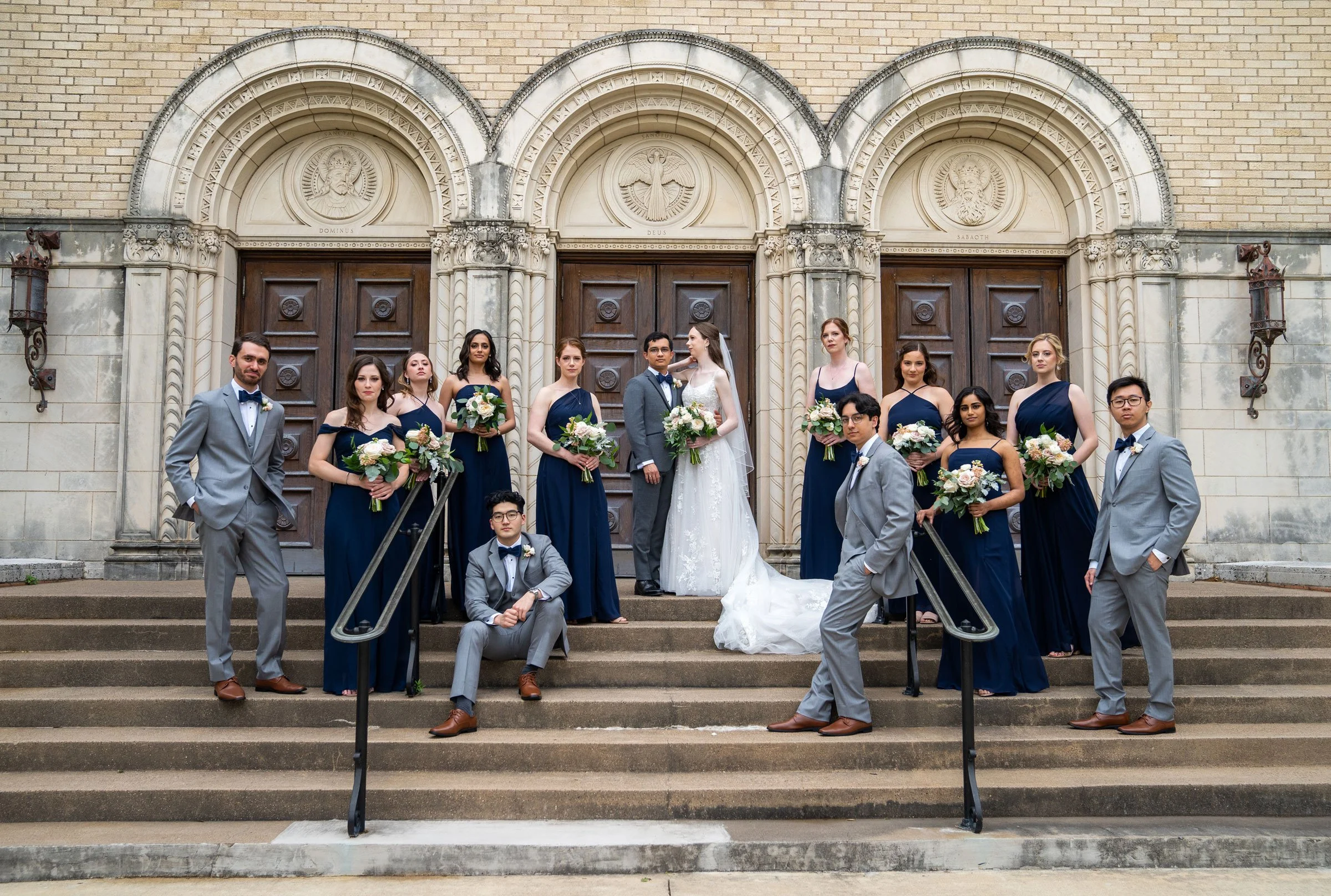 A group of people dressed in formal wedding attire, standing on the steps outside a church, with some holding bouquets of flowers, celebrating a wedding.