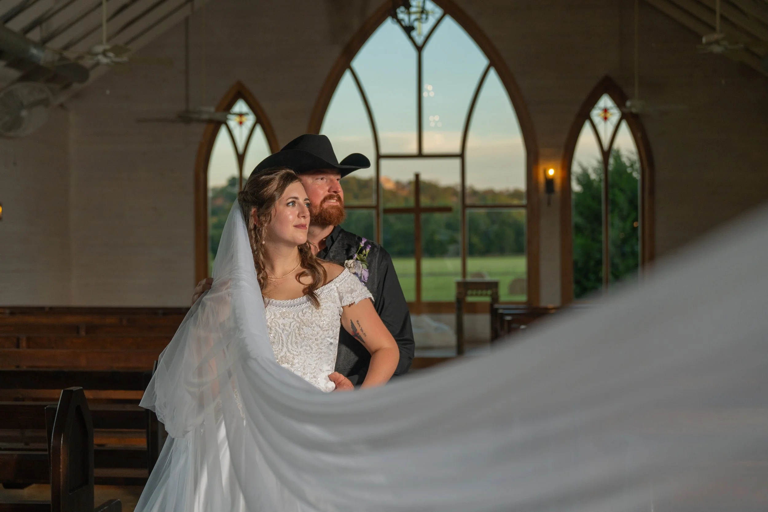 A bride and groom standing inside a church, with large arched windows and a scenic view outside. The bride is wearing a white lace wedding dress with a veil, and the groom is dressed in dark western attire with a black cowboy hat.